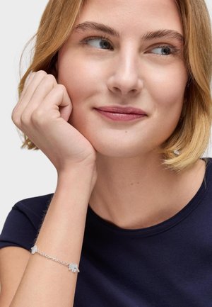 Bracciale d'argento con accentuazioni floreali su una delicata catena, indossato su un polso, abbinato a una maglietta blu navy.
