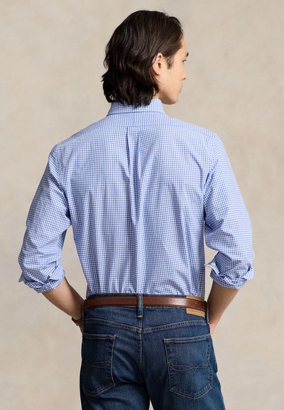 Mees, kes kannab helesinist ruudulist nööbitavat särki, mille varrukad on üles keeratud, tumedasiniseid teksaseid ja pruuni nahast vööd, seisab kaamera poole seljaga.