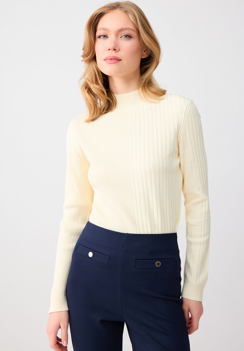 Blonde vrouw die een crèmekleurige geribbelde top met lange mouwen en hoge taille marineblauwe broek met knoopzakken draagt, staand tegen een witte achtergrond.