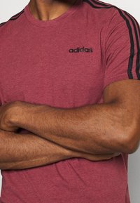 Camiseta roja jaspeada de manga corta, con el logo de adidas en negro y acentos de tres franjas negras a lo largo de los hombros. Textura de tela suave.