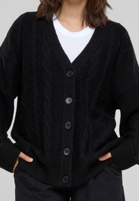 Cardigan de punto trenzado negro con escote en V y cinco grandes botones negros, terminado con puños y dobladillo acanalados para un toque de textura adicional.