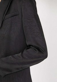 Blazer noir avec une texture froissée, doté d'une seule poche poitrine et d'une coupe ajustée. Le tissu a une apparence lisse et un subtil éclat.