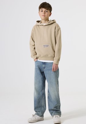 Sudadera beige con bolsillo frontal y texto azul, combinada con jeans sueltos de color azul claro y zapatillas blancas. Fondo neutro.