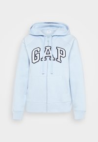Sudadera con cremallera de color azul claro, con bolsillos frontales, capucha con cordón y gran logo "GAP" en blanco y azul marino en el pecho.