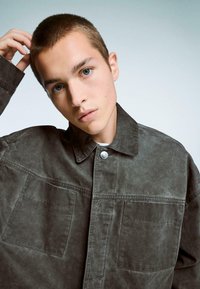 Temno zelena, strukturirana denim overshirt s koničastim ovratnikom, štirimi žepi in srebrnim gumbom na pritisni gumb na sprednji strani.