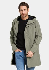 Manteau imperméable vert olive avec capuche, boutons-pression et doublure intérieure noire, porté sur un pull texturé noir et un jean bleu.