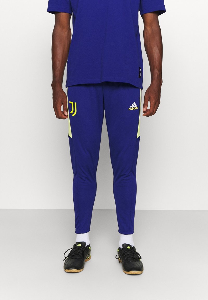 Mężczyzna w niebieskich spodniach dresowych Adidas Juventus z żółtymi logo i czarnych butach sportowych Adidas, stojący na jednolitym tle.