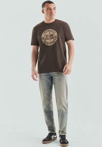 Brązowy bawełniany t-shirt z okrągłym graficznym wzorem tekstowym, w połączeniu z wyblakłymi niebieskimi dżinsami i czarnymi trampkami z jasnobrązową podeszwą.