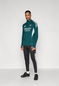 Performance trackjakke i blågrøn med hvide detaljer, lange ærmer og høj krave; sorte sportsbukser med hvide striber i siden og sorte sko.