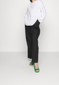 Top bianco a maniche lunghe con scollatura a chiave, abbinato a pantaloni neri a gamba larga e sandali verde con tacco a blocco e cinturini.