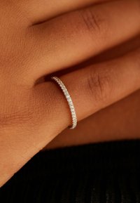 Dunne zilveren ring bezet met kleine, aaneengesloten ronde, heldere stenen, geplaatst op een hand met getextureerde huid, op een donkere, getextureerde ondergrond.