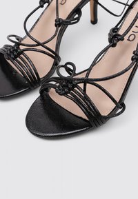 Sandalias de tacón alto negras con intrincadas correas anudadas, textura de cuero suave, diseño de punta abierta y un delicado tacón de aguja.