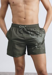 Olívazöld fürdőnadrág elasztikus derékrésszel, húzózsinórral, oldalzsebekkel és a bal oldalon jól látható logóval.