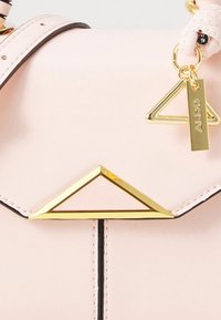 Vaaaleanpunainen keinonahkainen käsilaukku, jossa on kullanvärinen kolmion muotoinen koriste ja kullanvärinen nimilappu. Laukussa on kontrastivärinen musta reuna ja teksturoitu hihna.