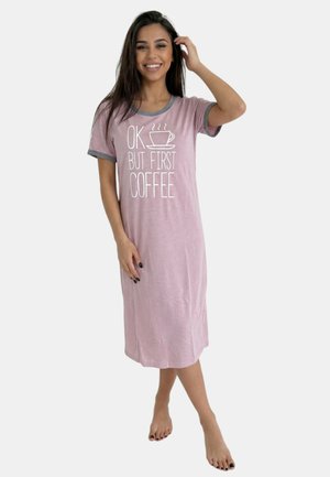 Mujer sonriente con cabello largo y oscuro, vistiendo un camisón rosa con ribete gris y gráfico de texto y taza, descalza.