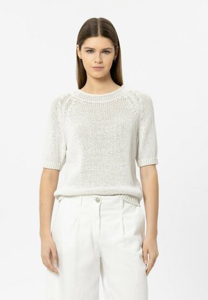 Young woman wearing a white knit short-sleeve sweater and high-waisted white pants, standing against a plain white background.