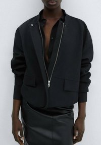 Veste bomber noire avec une fermeture éclair à l'avant, de grandes poches latérales et une coupe décontractée. Portée sur une chemise noire et une jupe en cuir, créant un look superposé.