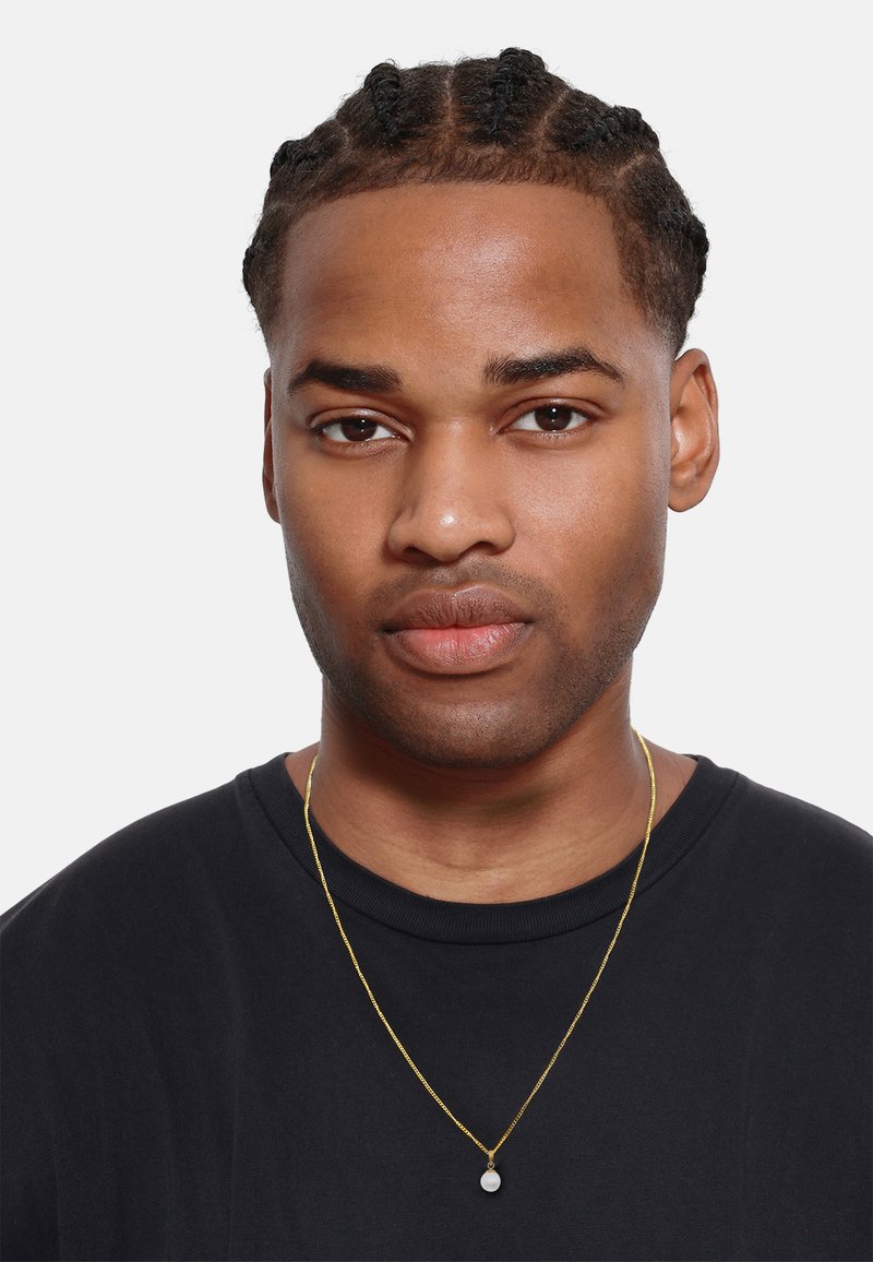 Guldkæde med en rund, hvid vedhæng, båret over en sort t-shirt. Håret er stylet i korte fletninger. Baggrunden er ensfarvet hvid.