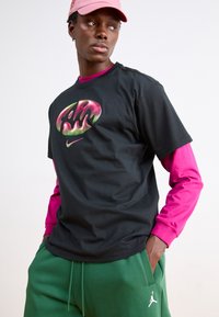 Svart t-shirt med en färgglad grafisk logotyp, bärs över en ljusrosa långärmad tröja, i kombination med gröna mjukisbyxor med en vit logotyp.