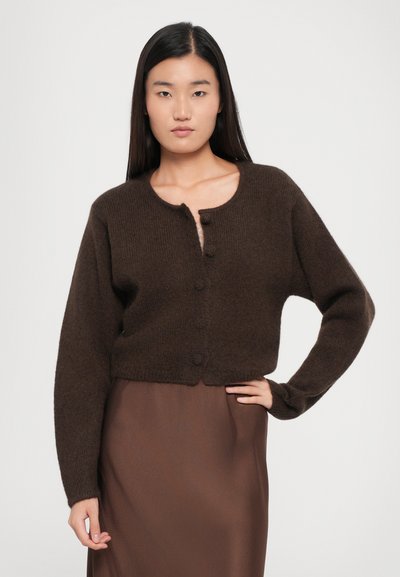 Brown cropped cardigan made of a soft material, featuring a round neck, five buttons, and long sleeves. Worn over a brown skirt.