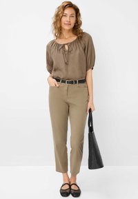 Blouse marron avec col à nouer, manches bouffantes ; pantalon kaki ; ceinture noire ; sac tissé noir ; chaussures noires avec motif découpé ; pantalon coupe droite.