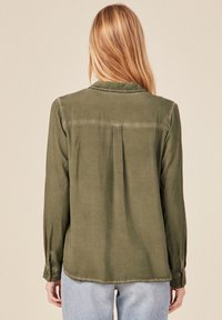 Camisa de manga larga verde oliva con un pliegue en la parte posterior, dobladillo curvado y puños abotonados, hecha de una tela suave y lisa.