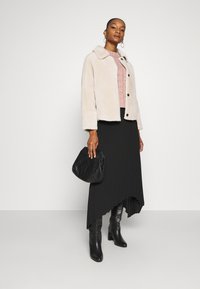 Cream faux fur jacket, pink cable-knit sweater, black asymmetric pleated skirt, tall black heeled boots, and a quilted black handbag.
