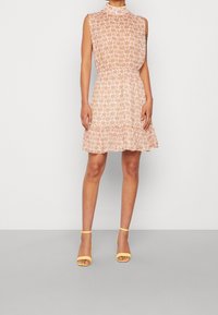 Robe courte sans manches de couleur rose clair avec un motif floral, taille froncée et ourlet volanté. Associée à des sandales à brides de cheville jaunes.