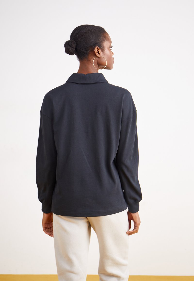 Polo negro de manga larga con cuello, que presenta un corte relajado y una textura suave. Diseño sencillo, combinado con pantalones de color claro.