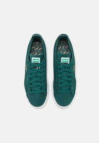 Sneakers verde in suede con suole in gomma bianca, caratterizzati da lacci piatti, colletto imbottito e un'etichetta con marchio sulla linguetta. Pattern testurizzato visibile sulla soletta.