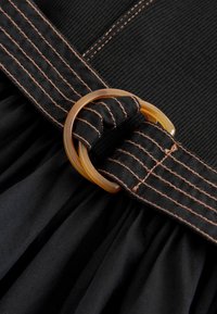 Gros plan d'une ceinture en tissu noir avec des coutures marron passant à travers une boucle ronde marron translucide sur un tissu noir plissé.