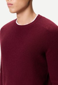 Vinröd stickad tröja med ribbad struktur, rund halsringning och åtsittande design. Närbild på axelsöm och tygdetalj.