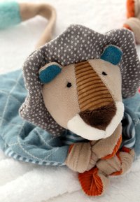 Jouet lion en tissu doux avec une crinière rayée, des oreilles bleues, et une couverture bleue à motifs enveloppée autour de son corps, allongé sur une surface blanche.