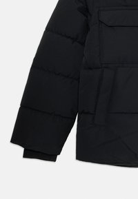 Manche de veste noire rembourrée avec un quilting horizontal, une poche à rabat rectangulaire au-dessus du poignet et un poignet côtelé visible à l'extrémité de la manche.