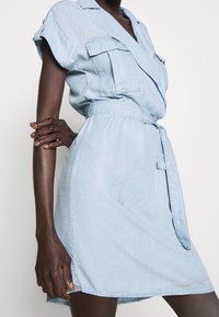 Jasnoniebieska dżinsowa sukienka z krótkimi rękawami, z kopertowym fasonem, dwiema kieszeniami na piersiach i przewiązywanym paskiem w talii. Gładka tekstura, długość do kolan.