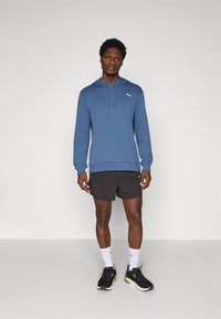Sudadera azul con capucha y cordón, pantalones cortos negros y zapatillas deportivas negras. Presenta un pequeño logo blanco en el pecho y calcetines blancos.