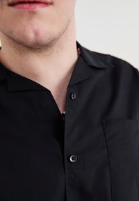 Svart kortermet skjorte med avslappet krage, frontlomme og to svarte knapper, laget av glatt stoff.