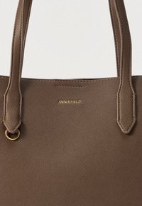 Barna texturált műbőrből készült válltáska, masszív fogantyúkkal és az elülső részén arany "ANNA FIELD" logóval.