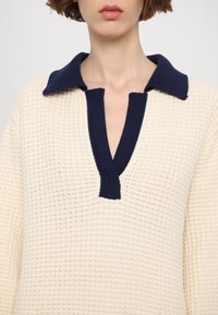 Femme portant un pull en maille texturée crème avec un col profond en V et un col et bord bleu marine.