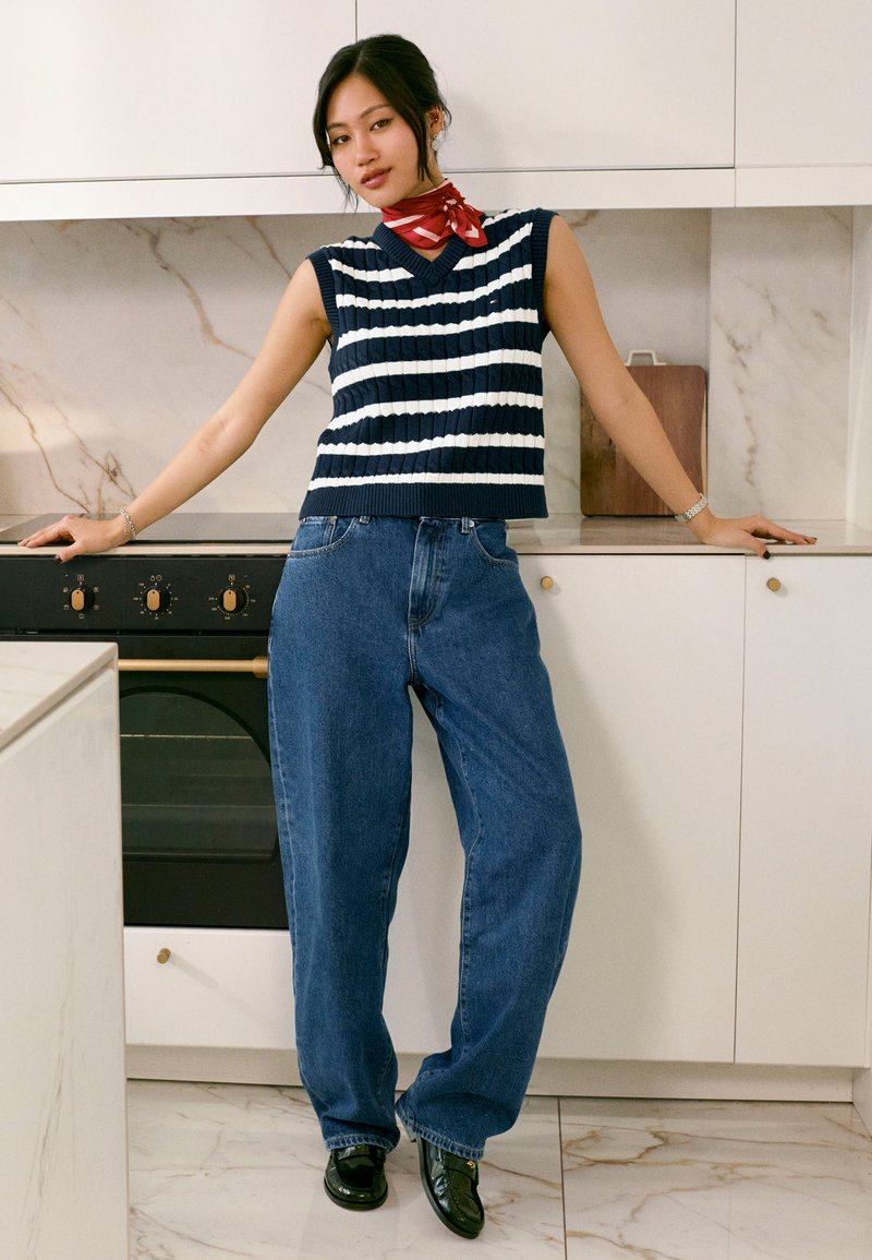 Jeune femme appuyée sur le plan de travail de la cuisine, portant un pull sans manches rayé bleu marine et blanc, un jean bleu ample, un foulard rouge autour du cou et des chaussures noires.
