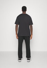 Schwarzes Baumwoll-T-Shirt mit kurzen Ärmeln, lockere Passform. Kombiniert mit schwarzen Hosen; der Träger steht vor einem hellgrauen Hintergrund.