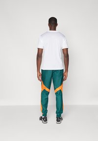 Chemise blanche à manches courtes et pantalon de jogging vert avec des accents orange. Le pantalon présente des poignets élastiques et des panneaux latéraux contrastants. Chaussures noires.