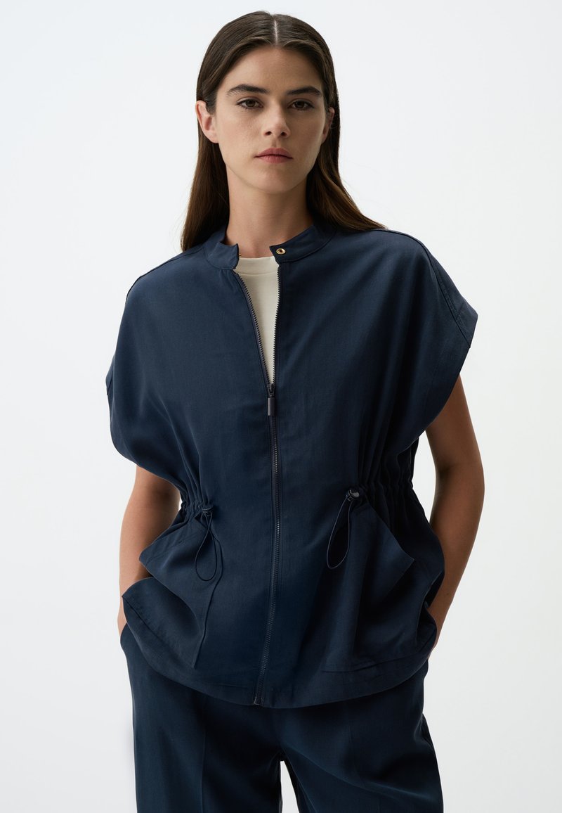 Veste courte bleu marine à manches courtes avec col montant, fermeture éclair sur le devant et taille ajustable par cordon, dotée de deux poches latérales et d'une texture de tissu lisse.