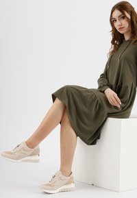 Abito verde oliva con cappuccio e vita arricciata; sneakers beige con dettagli in suede e tacco metallico. Tessuto liscio e vestibilità comoda.