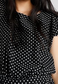 Blouse noire à pois blancs, coupe ample, tissu doux, col rond et manches courtes, avec un détail froncé à la taille.