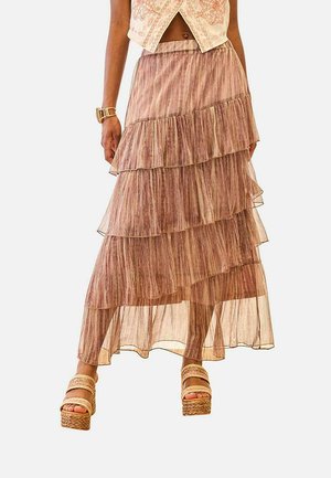 Mujer vistiendo una falda malva transparente de varios niveles con un top bordado corto y sandalias beige de plataforma, de pie frente a un fondo liso.