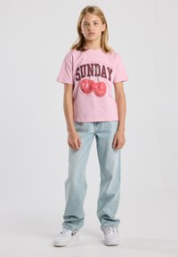 Camiseta de algodón rosa con mangas cortas, que presenta una impresión de "SUNDAY" y un gráfico de cerezas rojas. Combinada con unos vaqueros de denim azul claro y zapatillas blancas.
