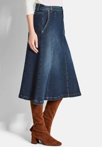 Jupe en denim évasée de couleur bleu foncé avec taille haute, poches latérales et coutures visibles ; associée à des bottes en daim marron montantes jusqu'aux genoux.