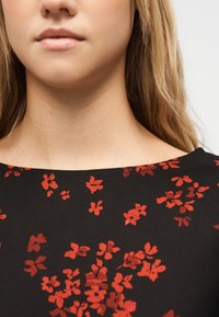 Gros plan sur le bas du visage, le cou et le haut de la poitrine d'une personne portant un haut noir à motif floral rouge.