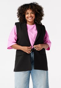 Glunderende vrouw met krullend haar, gekleed in een oversized roze shirt, een zwarte mouwloze blazer, lichtblauwe spijkerbroek en meerdere ringen tegen een witte achtergrond.